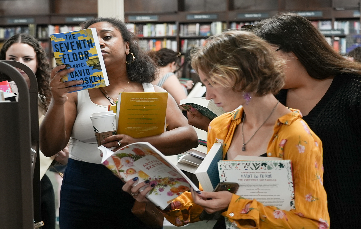 People browsing books in a bookstore, with shelves in the background. One person in a floral shirt holds a colorful book.