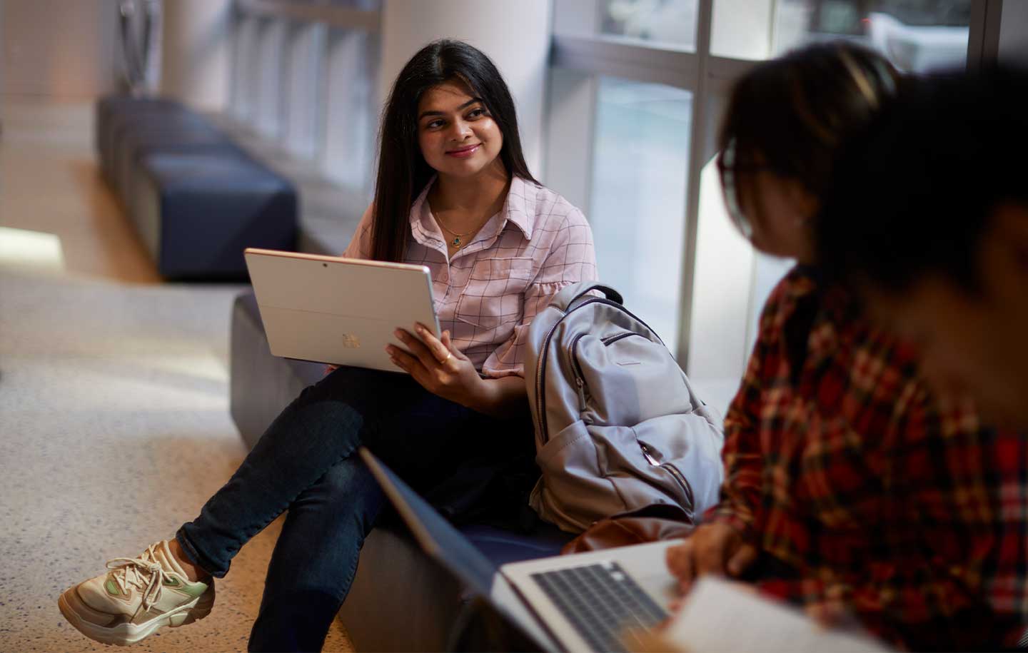 A woman in casual clothing, seated in a bright hallway, holds a laptop and smiles at a friend.