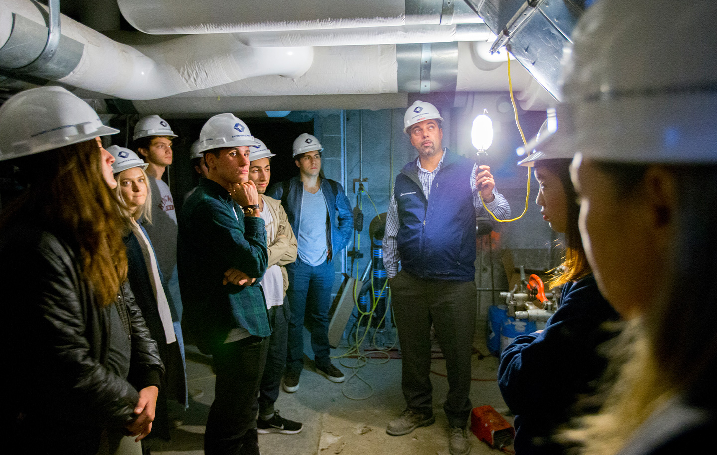 A group of people wearing hard hats stand in a dimly lit industrial basement. A man holds a light, explaining something.