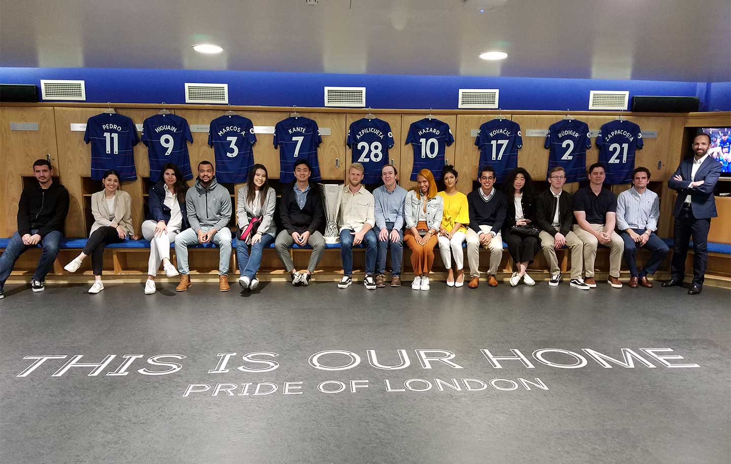 A group of people seated in a locker room beneath blue jerseys on a wooden rack. The floor reads "This is our home, Pride of London."
