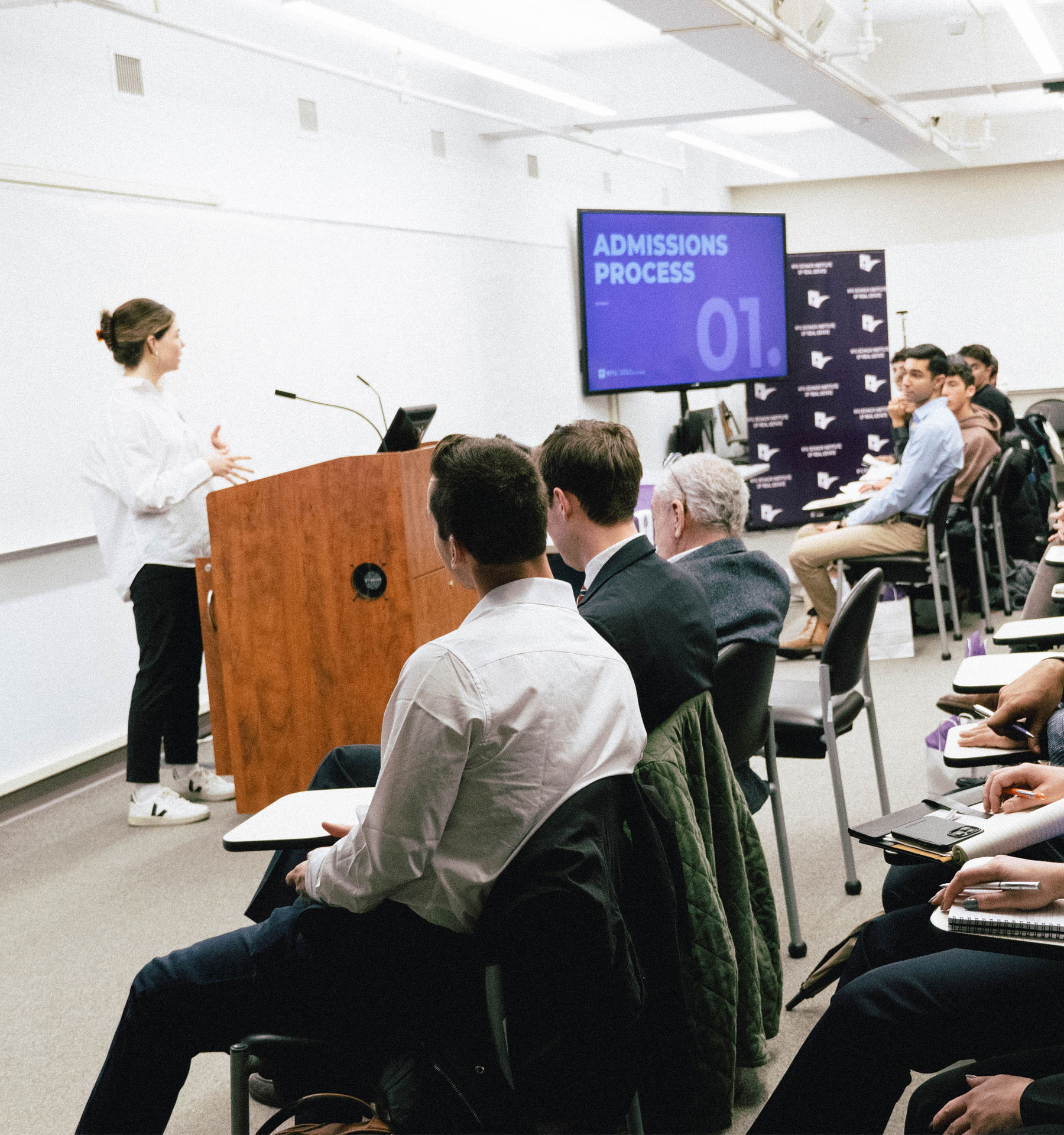 A student stands at a podium in a classroom presenting on the admissions process.
