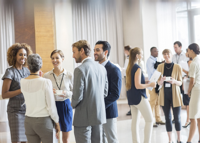 A crowd of professionals wearing nametags and holding information materials happily engage in small groups in a bright atrium.