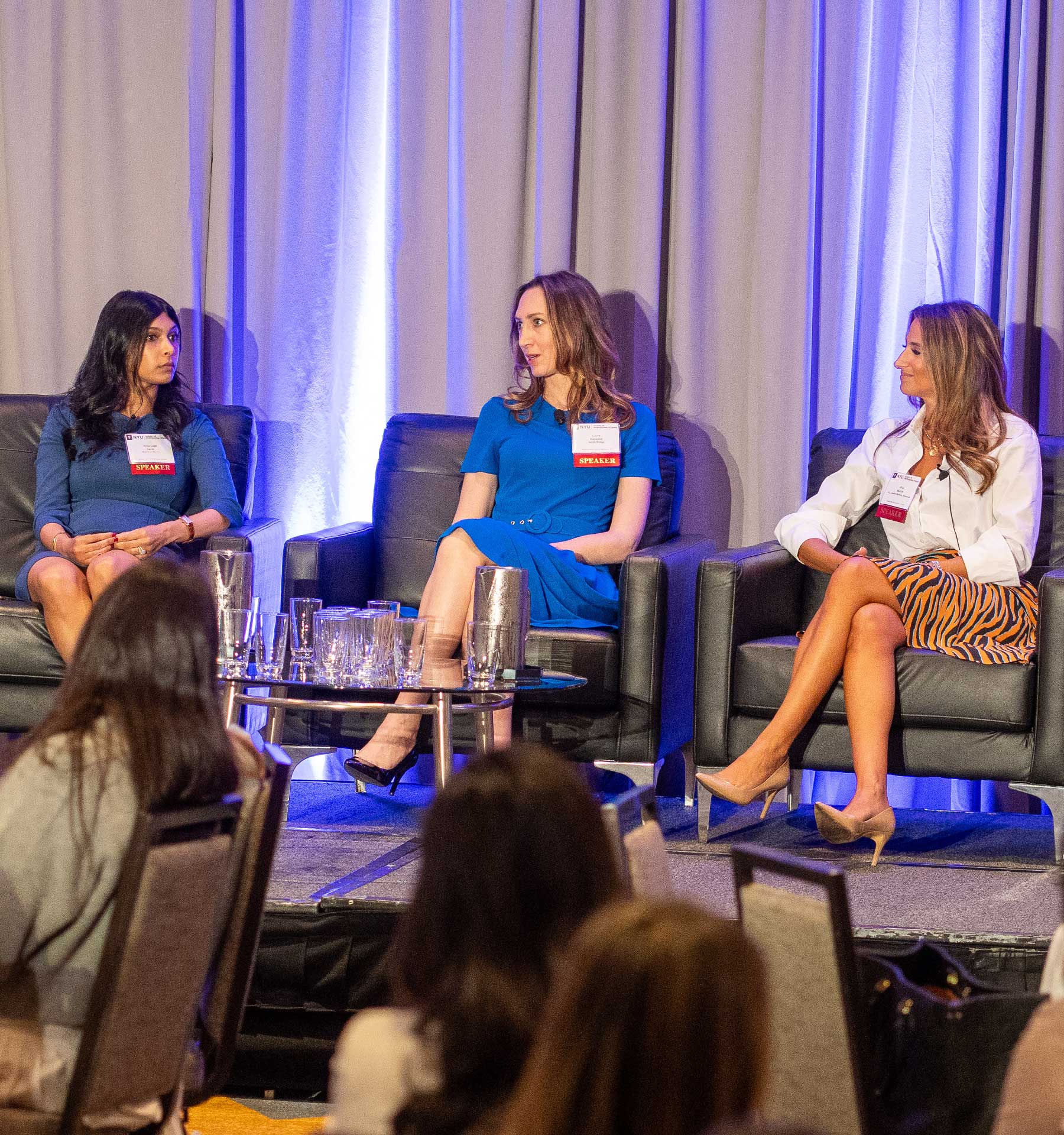 Three women sit on a panel in a conference room, engaged in discussion. They are seated on black armchairs with a table of glasses in front, under soft blue lighting. The audience listens attentively.