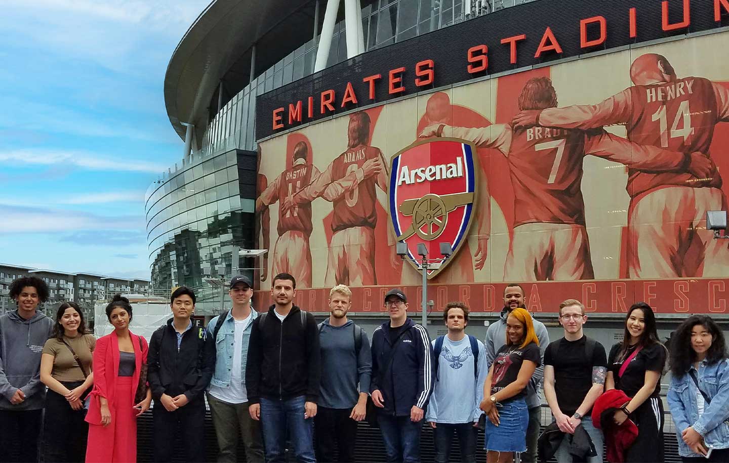 A group of people stands in front of the Emirates Stadium with an Arsenal mural and logo in the background.
