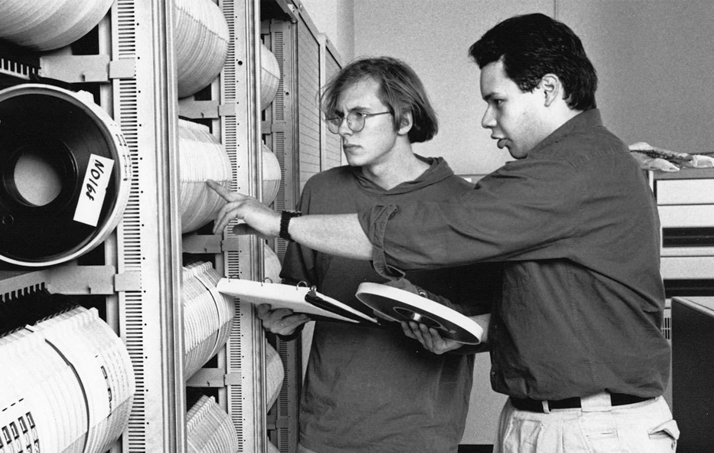 Two individuals examining magnetic tape reels in a data storage room.