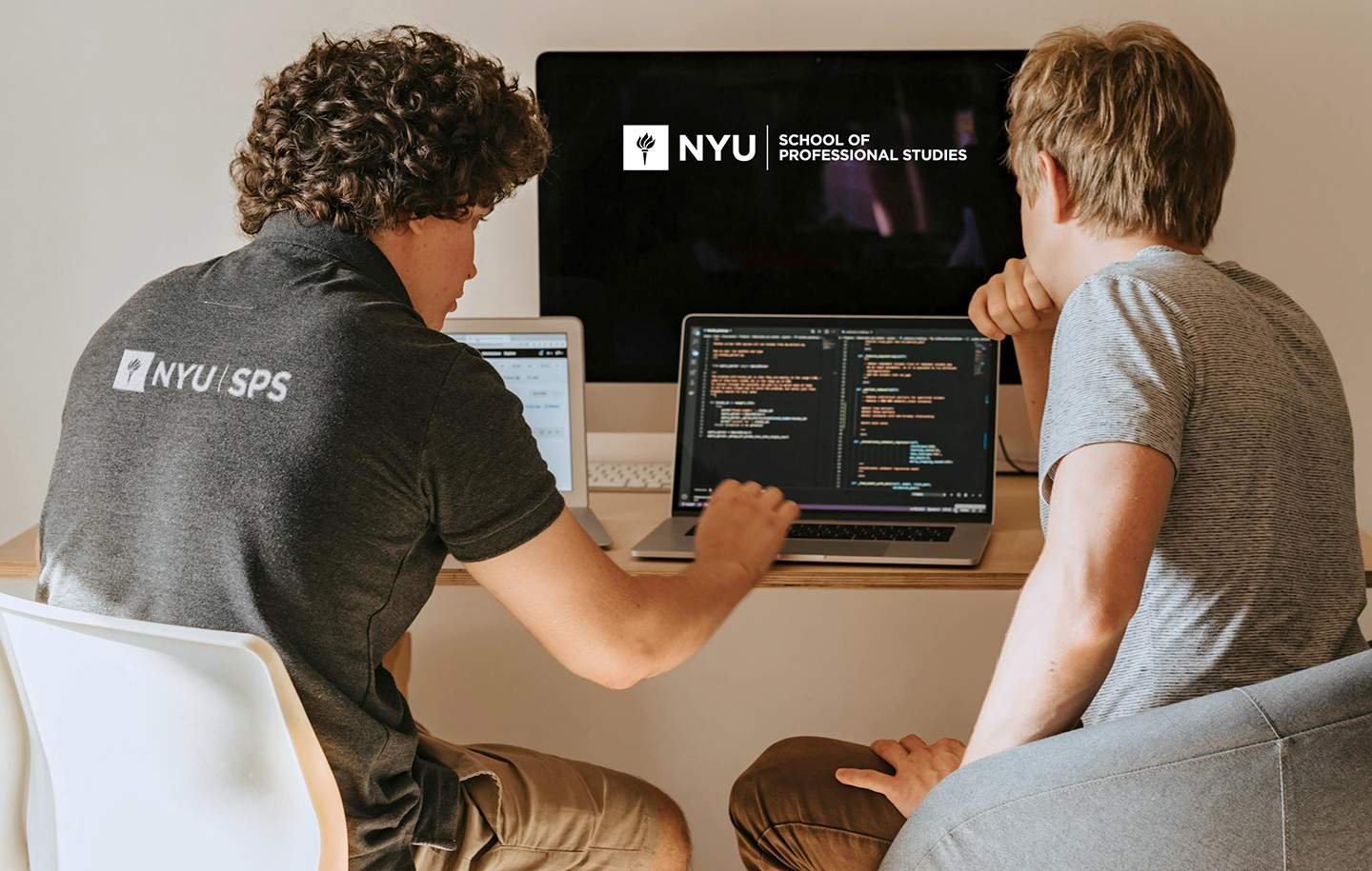 Two NYU SPS students sit at a desk with a computer and two laptops, one of which has lines of code