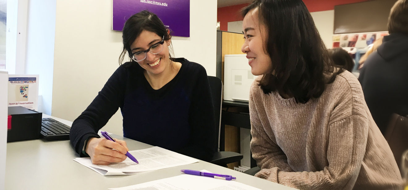 An NYU SPS student sits at a table in an academic advising office with her advisor who holds a pen to paper and both are smiling