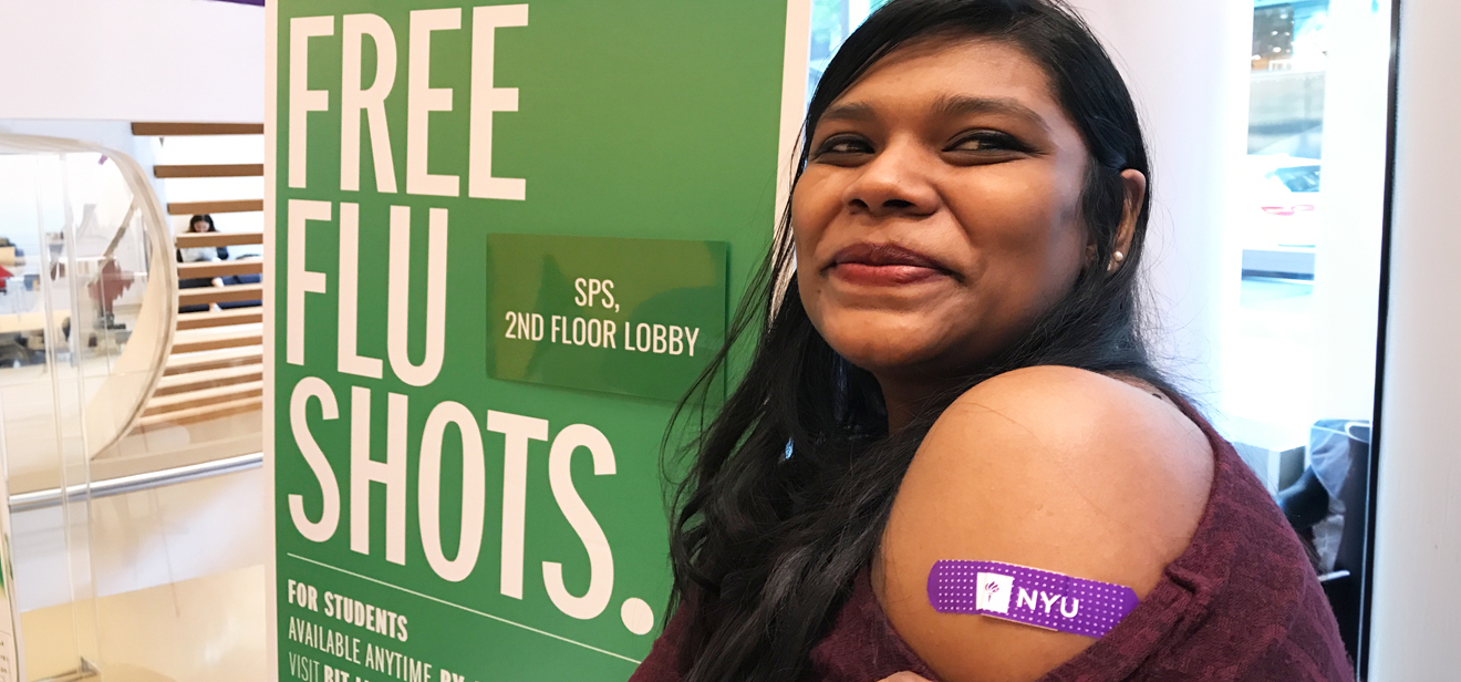 NYU SPS student softly smiles with a purple NYU bandage on her upper arm in front of a green free flu shots sign