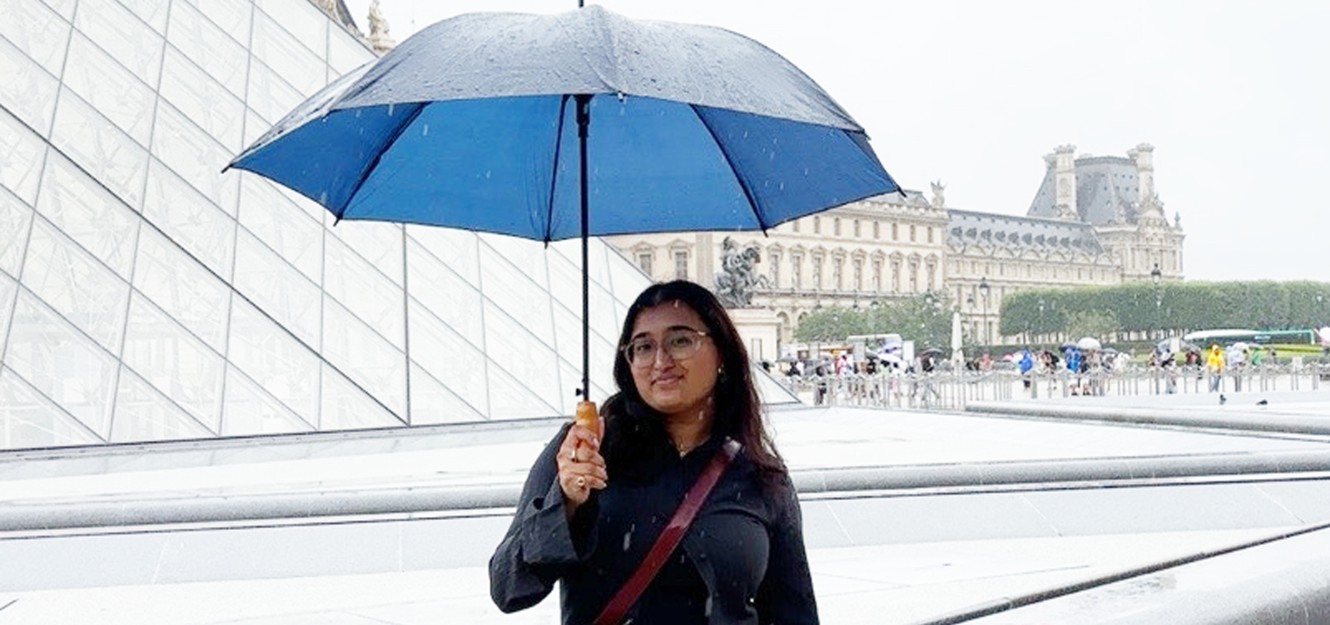 Meahjabeen Hoque smiles and holds a blue umbrella in front of the Louvre in Paris
